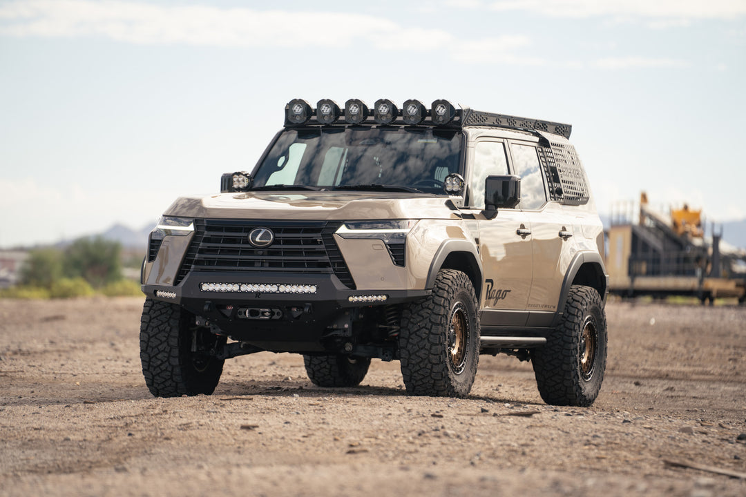 A heavily modified Lexus GX 550 SUV in a desert sand color, equipped with a Rago roof-rack, front bumper, and molle panels, parked on a dirt ground.