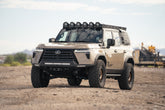A heavily modified Lexus GX 550 SUV in a desert sand color, equipped with a Rago roof-rack, front bumper, and molle panels, parked on a dirt ground.