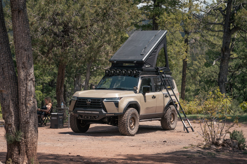 GX 550 fully equipped at camp with off road bumpers, roof rack, tent, light brackets, and molle panels. Roof top tent is opened with ladder extending down.