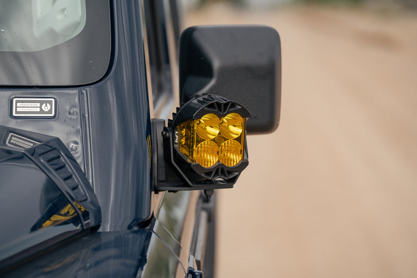 A close-up shot of Ditch Light Brackets for the 2022-2026 INEOS Grenadier & Quartermaster with auxiliary light mounted on the side mirror, with a blurred sandy road in the background.